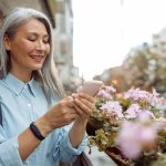Smiling grey haired Asian woman uses cellphone standing on outdoors terrace